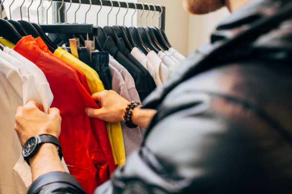 A customer, focused on browsing a clothing rack, sorts through various colorful shirts in shades of red, white, and yellow. With a watch and bracelet adorning their wrist, the priority is clearly on exploring the vibrant clothes rather than drawing attention to their face.